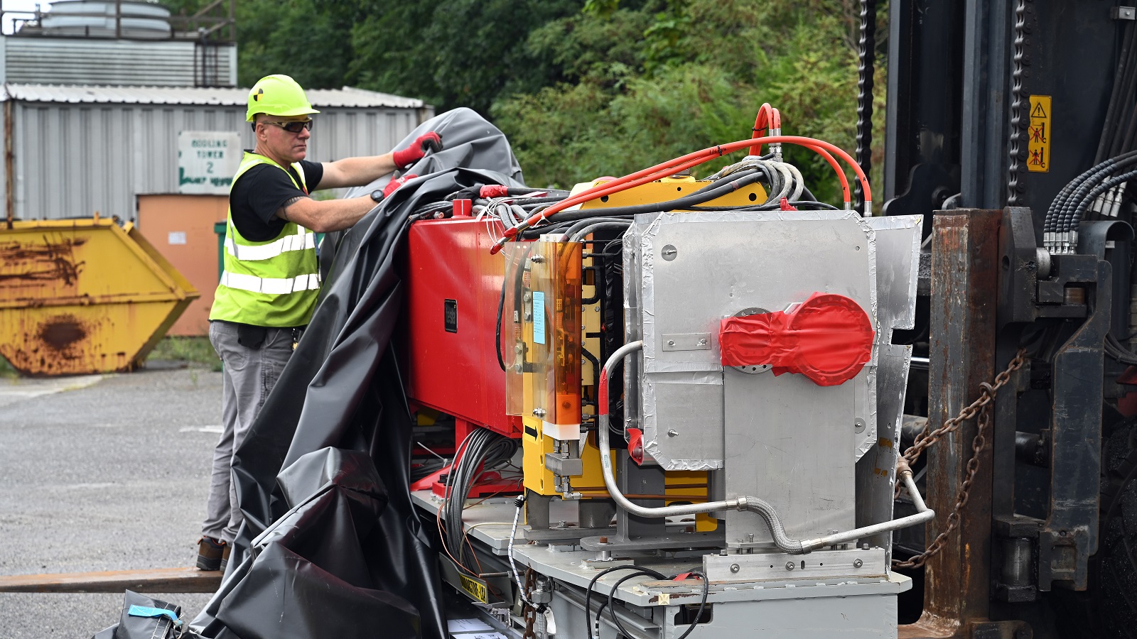 A worker in a safety vest at Brookhaven National Laboratory removes the black tarp from a module of magnets and wires.