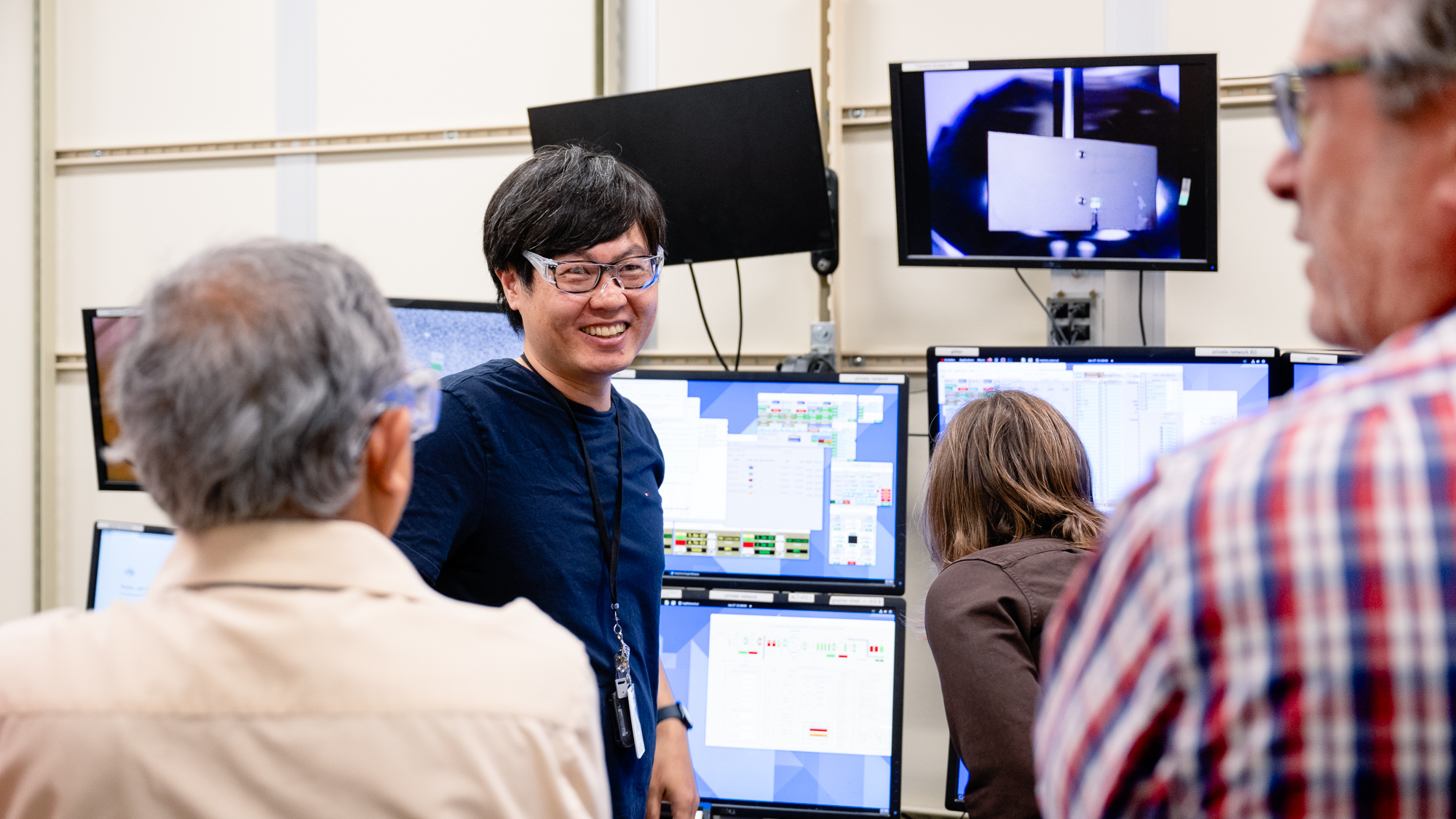 A man smiles near a monitor showing a grey card with a spot of white light.