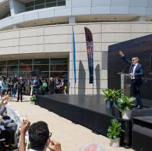 : A man in a suit holds aloft a glass with sparkling cider, and a crowd of people join him with their own glasses. 